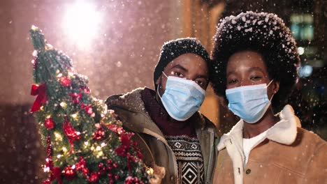 Close-up-view-of-joyful-African-American-couple-wearing-facial-masks-holding-a-Christmas-tree-and-smiling-at-camera-while-it¬¥s-snowing-on-the-street-in-Christmas