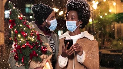 Nahaufnahme-Eines-Fröhlichen-Afroamerikanischen-Paares-Mit-Gesichtsmasken,-Das-Spricht-Und-Etwas-Am-Telefon-Beobachtet,-Während-Es-Zu-Weihnachten-Auf-Der-Straße-Schneit