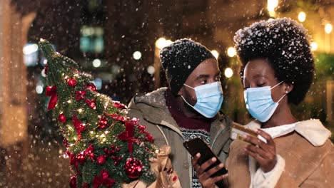 Nahaufnahme-Eines-Fröhlichen-Afroamerikanischen-Paares-Mit-Gesichtsmasken,-Das-Online-Mit-Dem-Smartphone-Redet-Und-Einkauft,-Während-Es-Zu-Weihnachten-Auf-Der-Straße-Schneit