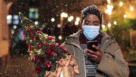 Porträt-Eines-Jungen-Afroamerikanischen-Mannes-Mit-Gesichtsmaske,-Der-Auf-Dem-Smartphone-Tippt-Und-Einen-Weihnachtsbaum-Auf-Der-Straße-Hält,-Während-Es-Zu-Weihnachten-Schneit