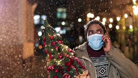 Portrait-of-young-african-american-man-wearing-facial-mask-talking-on-smartphone-and-holding-a-christmas-tree-on-the-street-while-it¬¥s-snowing-in-Christmas