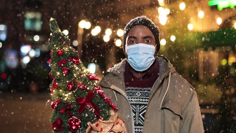 Porträt-Eines-Jungen-Afroamerikanischen-Mannes-Mit-Gesichtsmaske,-Der-Einen-Weihnachtsbaum-Hält-Und-Auf-Der-Straße-In-Die-Kamera-Lächelt,-Während-Es-Zu-Weihnachten-Schneit