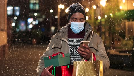 Portrait-of-young-african-american-man-wearing-facial-mask-typing-on-smartphone-and-holding-shopping-bags-on-the-street-while-it¬¥s-snowing-in-Christmas