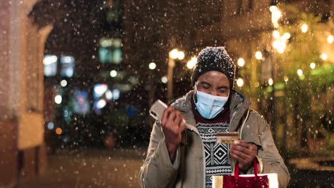 Porträt-Eines-Jungen-Afroamerikaners-Mit-Gesichtsmaske,-Der-Auf-Dem-Smartphone-Tippt-Und-Online-Auf-Der-Straße-Einkauft,-Während-Es-In-Weihnachten-Schneit