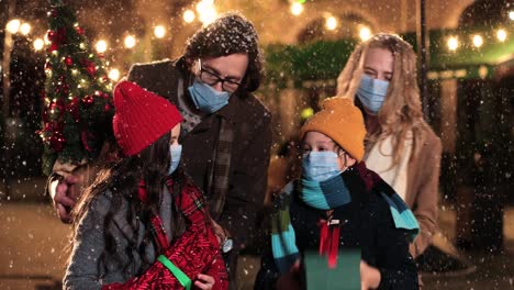 Nahaufnahme-Einer-Kaukasischen-Glücklichen-Familie,-Die-Gesichtsmasken-Trägt-Und-Geschenke-Auf-Der-Straße-Hält,-Während-Es-In-Weihnachten-Schneit