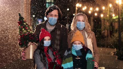 Close-up-view-of-Caucasian-happy-family-wearing-facial-masks-holding-presents-and-smiling-at-camera-on-the-street-while-it¬¥s-snowing-in-Christmas