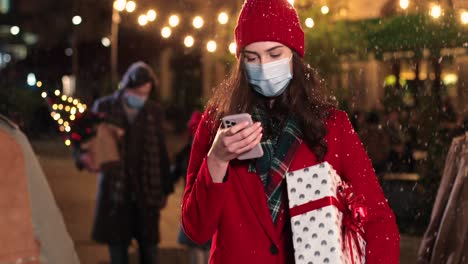 Portrait-of-happy-beautiful-young-woman-typing-on-smartphone-and-walking-down-the-street-while-it¬¥s-snowing-in-Christmas