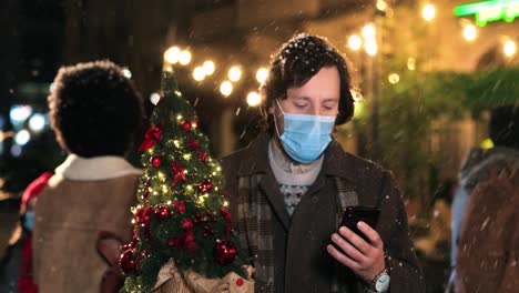 Portrait-of-caucasian-man-typing-on-smartphone-and-holding-a-christmas-tree-on-the-street-while-it¬¥s-snowing-in-Christmas