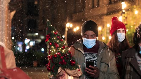 Portrait-of-african-american-man-typing-on-smartphone-and-holding-a-christmas-tree-on-the-street-while-it¬¥s-snowing-in-Christmas