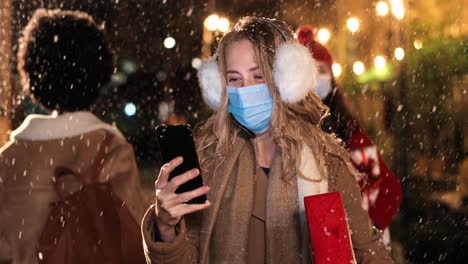 Vista-De-Cerca-De-Una-Joven-Feliz-Y-Hermosa-Con-Orejeras-Y-Máscara-Facial-Escribiendo-En-Un-Smartphone-En-La-Calle-Mientras-Nieva-En-Navidad