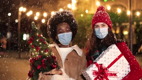 Glückliche-Kaukasische-Und-Afroamerikanische-Freundinnen,-Die-Geschenke-Halten-Und-Auf-Der-Straße-In-Die-Kamera-Lächeln,-Während-Es-In-Weihnachten-Schneit