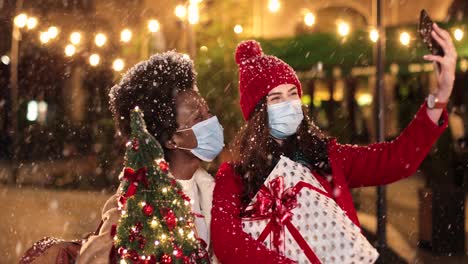 Glückliche-Kaukasische-Und-Afroamerikanische-Freundinnen,-Die-Ein-Selfie-Machen-Und-Geschenke-Auf-Der-Straße-Halten,-Während-Es-In-Weihnachten-Schneit