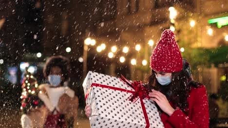 Nahaufnahme-Einer-Jungen-Kaukasischen-Frau-Mit-Medizinischer-Maske,-Die-Eingepackte-Geschenkbox-Schüttelt-Und-Versucht-Zu-Erraten,-Was-Auf-Der-Straße-Ist,-Während-Es-Zu-Weihnachten-Schneit