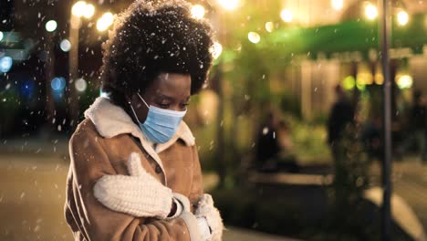Nahaufnahme-Einer-Afroamerikanischen-Frau-Mit-Gesichtsmaske,-Die-Erkältet-Ist-Und-Versucht,-Sich-Auf-Der-Straße-Aufzuwärmen,-Während-Es-Zu-Weihnachten-Schneit
