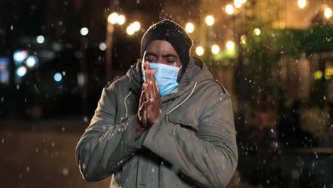 Nahaufnahme-Eines-Afroamerikanischen-Mannes-Mit-Gesichtsmaske,-Der-Erkältet-Ist-Und-Versucht,-Sich-Auf-Der-Straße-Aufzuwärmen,-Während-Es-Zu-Weihnachten-Schneit