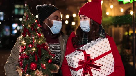 Nahaufnahme-Eines-Glücklichen-Multiethnischen-Paares,-Das-Gesichtsmasken-Trägt-Und-Weihnachtsgeschenke-Auf-Der-Straße-Hält,-Während-Es-Zu-Weihnachten-Schneit