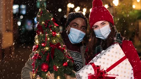 Nahaufnahme-Eines-Glücklichen-Multiethnischen-Paares,-Das-Gesichtsmasken-Trägt-Und-Weihnachtsgeschenke-Hält,-Während-Es-Auf-Der-Straße-In-Die-Kamera-Lächelt,-Während-Es-Zu-Weihnachten-Schneit