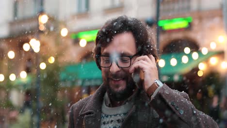 Nahaufnahme-Eines-Kaukasischen-Mannes-Mit-Brille,-Der-Auf-Der-Straße-Mit-Dem-Smartphone-Spricht,-Während-Es-In-Weihnachten-Schneit