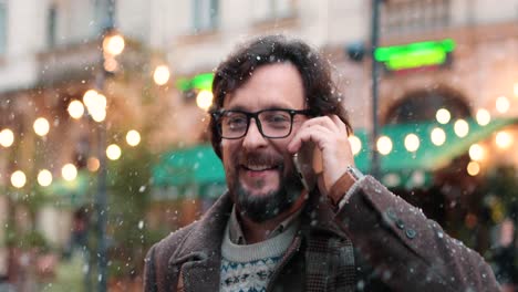 Nahaufnahme-Eines-Kaukasischen-Mannes-Mit-Brille,-Der-Auf-Der-Straße-Mit-Dem-Smartphone-Spricht,-Während-Es-In-Weihnachten-Schneit