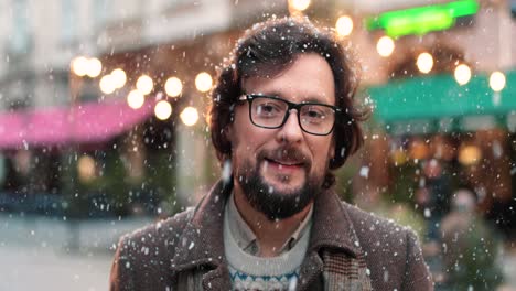 Nahaufnahme-Eines-Kaukasischen-Mannes-Mit-Brille,-Der-Auf-Der-Straße-In-Die-Kamera-Blickt,-Während-Es-Zu-Weihnachten-Schneit