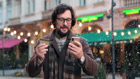 Nahaufnahme-Eines-Kaukasischen-Mannes-Mit-Brille,-Der-Online-Mit-Dem-Smartphone-Auf-Der-Straße-Einkauft,-Während-Es-In-Weihnachten-Schneit