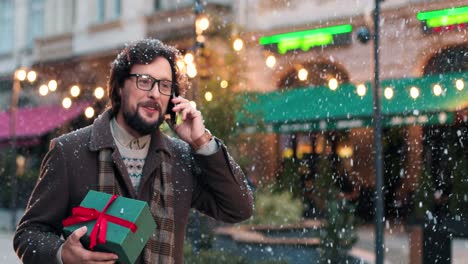 Vista-De-Cerca-De-Un-Hombre-Caucásico-Con-Anteojos-Sosteniendo-Un-Regalo-Y-Hablando-Por-Teléfono-Inteligente-En-La-Calle-Mientras-Nieva-En-Navidad