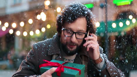 Nahaufnahme-Eines-Kaukasischen-Mannes-Mit-Brille,-Der-Ein-Geschenk-In-Der-Hand-Hält-Und-Auf-Der-Straße-Mit-Dem-Smartphone-Spricht,-Während-Es-Zu-Weihnachten-Schneit
