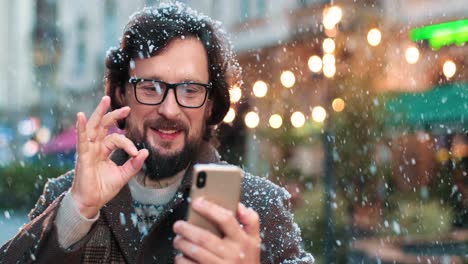 Nahaufnahme-Eines-Kaukasischen-Mannes-Mit-Brille,-Der-Einen-Videoanruf-Mit-Smartphone-Auf-Der-Straße-Tätigt,-Während-Es-In-Weihnachten-Schneit
