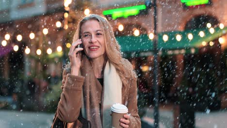 Vista-Cercana-De-Una-Mujer-Caucásica-Hablando-Por-Teléfono-Inteligente-En-La-Calle-Mientras-Nieva-En-Navidad