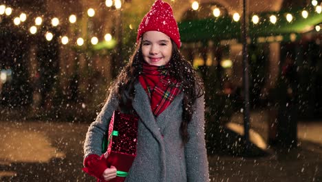 Portrait-of-happy-cute-girl-holding-christmas-present-and-smiling-at-camera-on-the-street-while-it‚Äôs-snowing-in-Christmas