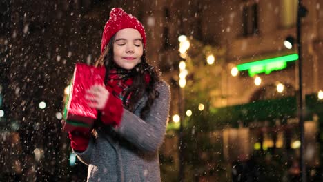 Portrait-of-happy-cute-girl-holding-christmas-present-and-smiling-at-camera-on-the-street-while-it‚Äôs-snowing-in-Christmas