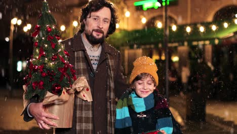 Portrait-of-father-and-his-little-son-holding-present-and-christmas-tree,-and-looking-at-camera-on-the-street-while-it‚Äôs-snowing-in-Christmas