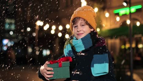 Portrait-of-happy-cute-boy-holding-christmas-present-and-smiling-at-camera-on-the-street-while-it‚Äôs-snowing-in-Christmas