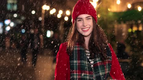 Close-up-view-of-caucasian-woman-in-red-coat-holding-a-present-and-smiling-at-camera-on-the-street-while-it‚Äôs-snowing-in-Christmas