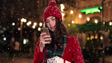 Vista-De-Cerca-De-Una-Mujer-Caucásica-Con-Abrigo-Rojo-Sosteniendo-Un-Regalo-Y-Usando-Un-Smartphone-En-La-Calle-Mientras-Nieva-En-Navidad