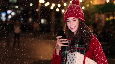 Nahaufnahme-Einer-Kaukasischen-Frau-Im-Roten-Mantel,-Die-Ein-Geschenk-Hält-Und-Ein-Smartphone-Auf-Der-Straße-Benutzt,-Während-Es-Zu-Weihnachten-Schneit