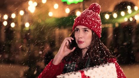 Vista-De-Cerca-De-Una-Mujer-Caucásica-Con-Abrigo-Rojo-Sosteniendo-Un-Regalo-Y-Hablando-Por-Teléfono-Inteligente-En-La-Calle-Mientras-Nieva-En-Navidad