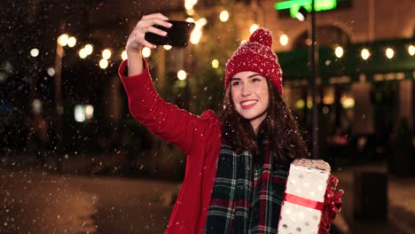 Nahaufnahme-Einer-Kaukasischen-Frau-Im-Roten-Mantel,-Die-Ein-Geschenk-Hält-Und-Ein-Selfie-Mit-Dem-Smartphone-Auf-Der-Straße-Macht,-Während-Es-Zu-Weihnachten-Schneit