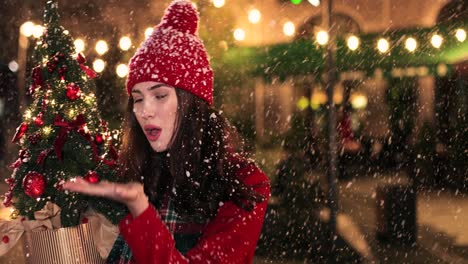 Close-up-view-of-caucasian-woman-in-red-coat-holding-a-present-and-christmas-tree,-trying-to-catch-the-snow-on-the-street-in-Christmas