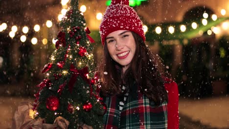 Nahaufnahme-Einer-Kaukasischen-Frau-Im-Roten-Mantel,-Die-Einen-Weihnachtsbaum-Hält-Und-Auf-Der-Straße-In-Die-Kamera-Lächelt,-Während-Es-Zu-Weihnachten-Schneit