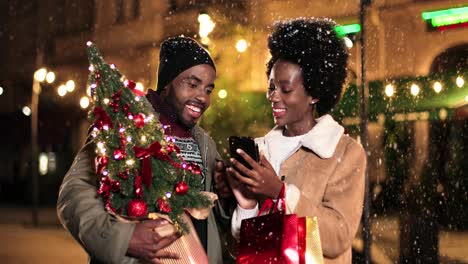 Nahaufnahme-Eines-Fröhlichen-Afroamerikanischen-Paares,-Das-Sich-Unterhält-Und-Etwas-Am-Telefon-Ansieht,-Während-Es-Zu-Weihnachten-Auf-Der-Straße-Schneit