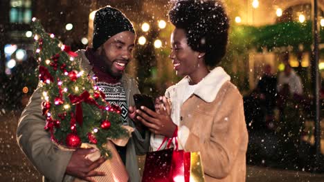 Nahaufnahme-Eines-Fröhlichen-Afroamerikanischen-Paares,-Das-Sich-Unterhält-Und-Etwas-Am-Telefon-Ansieht,-Während-Es-Zu-Weihnachten-Auf-Der-Straße-Schneit