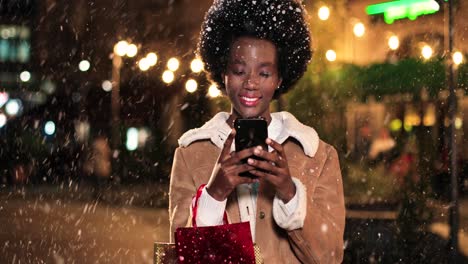 Close-up-view-of-african-american-woman-holding-shopping-bag-and-using-smartphone-on-the-street-while-it‚Äôs-snowing-in-Christmas