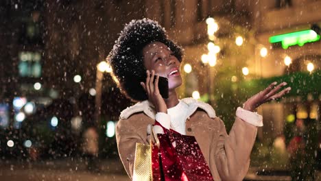 Nahaufnahme-Einer-Afrikanisch-amerikanischen-Frau,-Die-Eine-Einkaufstasche-Hält-Und-Auf-Der-Straße-Mit-Dem-Smartphone-Spricht,-Während-Es-In-Weihnachten-Schneit