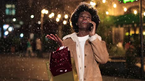 Vista-Cercana-De-Una-Mujer-Afroamericana-Sosteniendo-Una-Bolsa-De-Compras-Y-Hablando-Por-Teléfono-Inteligente-En-La-Calle-Mientras-Nieva-En-Navidad