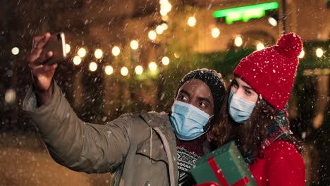 Vista-De-Cerca-De-Una-Alegre-Pareja-Multiétnica-Haciendo-Un-Selfie-Con-Un-Smartphone-Mientras-Nieva-En-La-Calle-En-Navidad