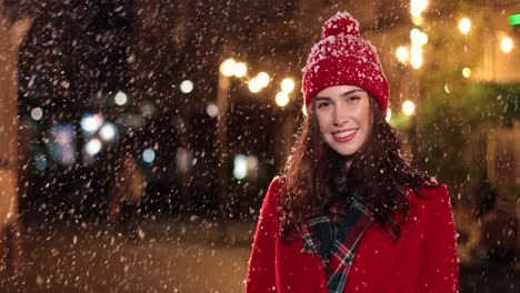 Cheerful-young-woman-in-red-coat-smiling-at-camera-while-it¬¥s-snowing-in-Christmas