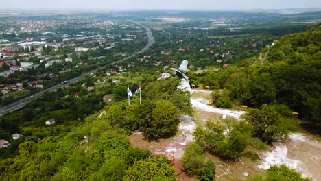 Flying-over-Tatabánya-city-in-Hungary,-turul-bird-statue-and-Hungarian-flags-view