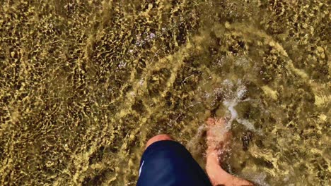 Caucasian-male-legs-and-feet-walking-through-ocean-waves-barefoot-towards-sandy-beach