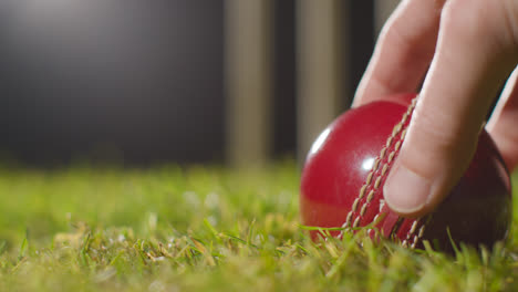 Cricket-Stillleben-Mit-Nahaufnahme-Einer-Hand,-Die-Einen-Ball-Aufhebt,-Der-Im-Gras-Vor-Stümpfen-Liegt